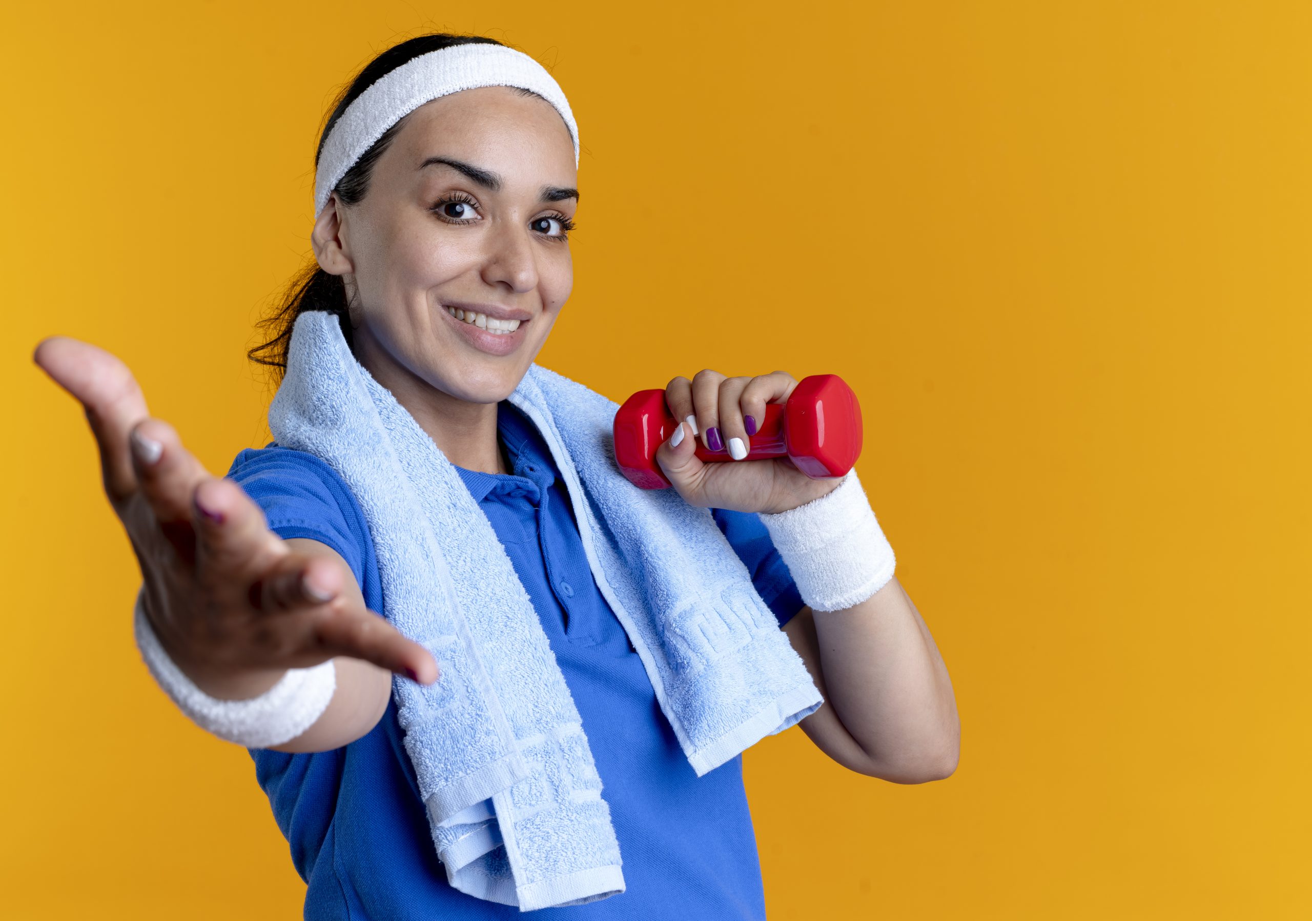 young smiling caucasian sporty woman wearing headband and wristbands with towel on neck holds dumbbell and holds hand out isolated on orange background with copy space